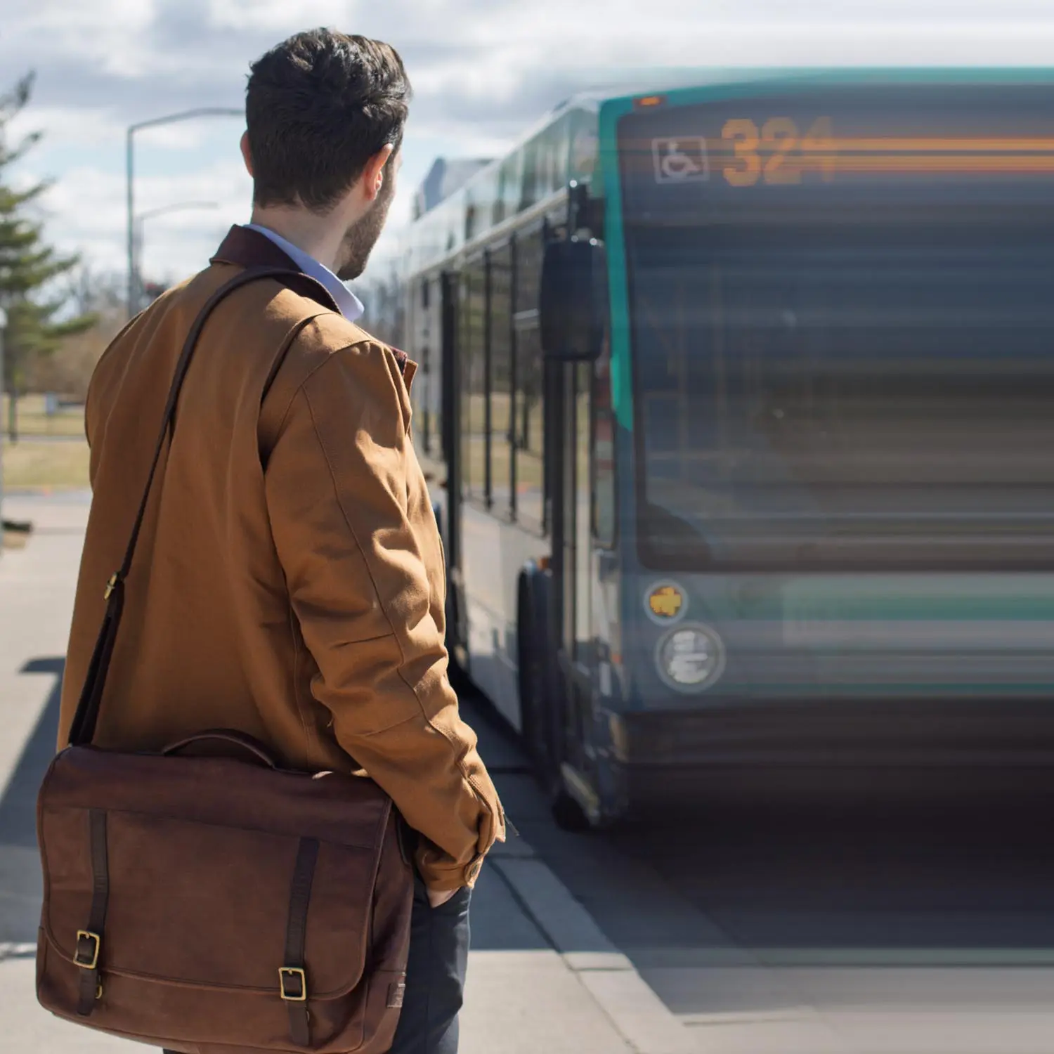 Man with a brown jacket and leather messenger bag waiting at a bus stop as a bus approaches.