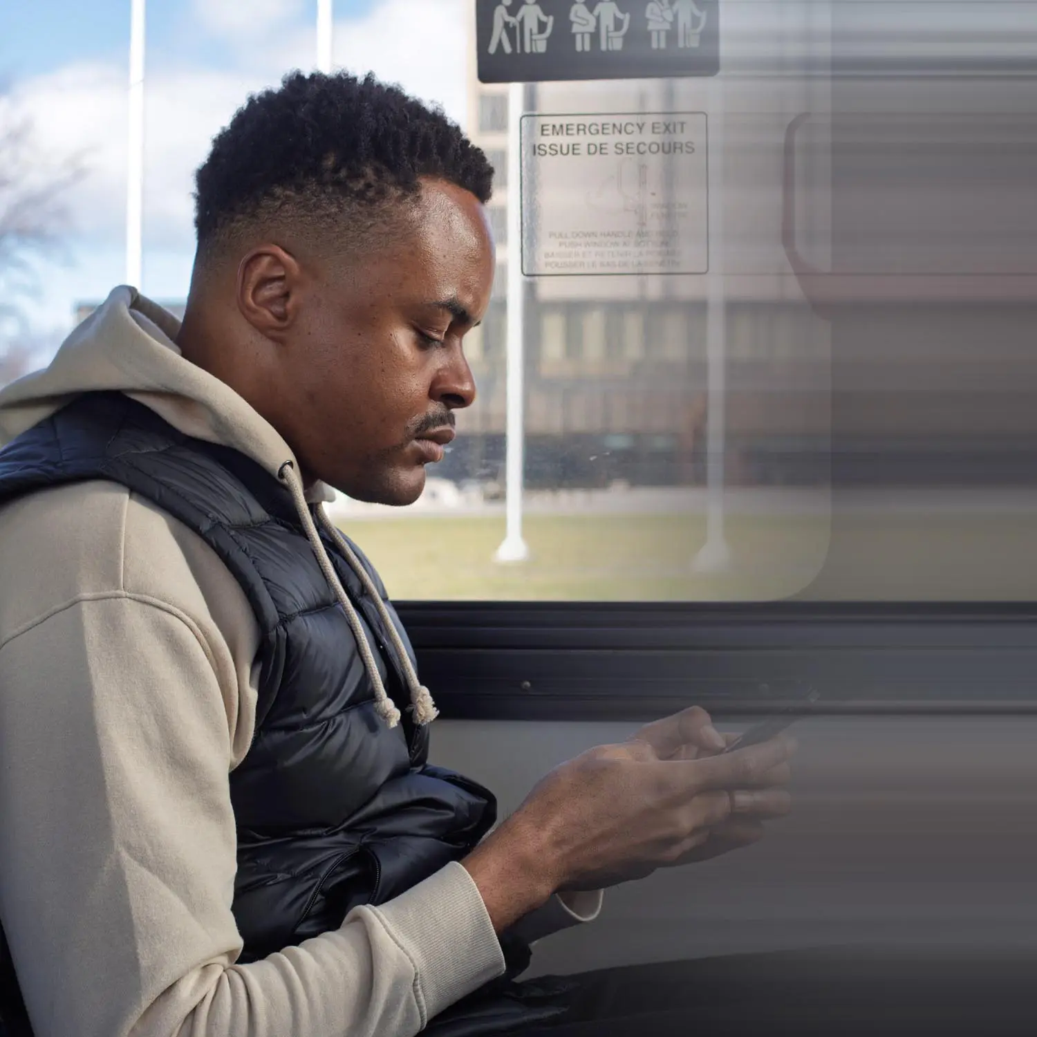 Man wearing a beige hoodie and black puffer vest sitting by a bus window, looking at his phone.