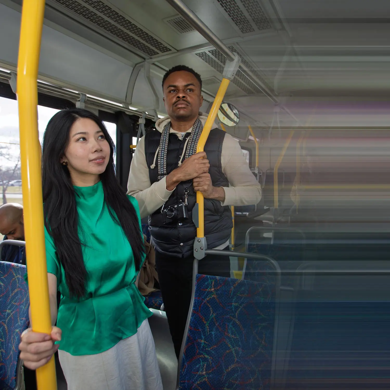 Two people standing inside a bus, holding yellow poles, with one woman wearing a green blouse and a man wearing a black vest and beige hoodie with a camera around his neck.