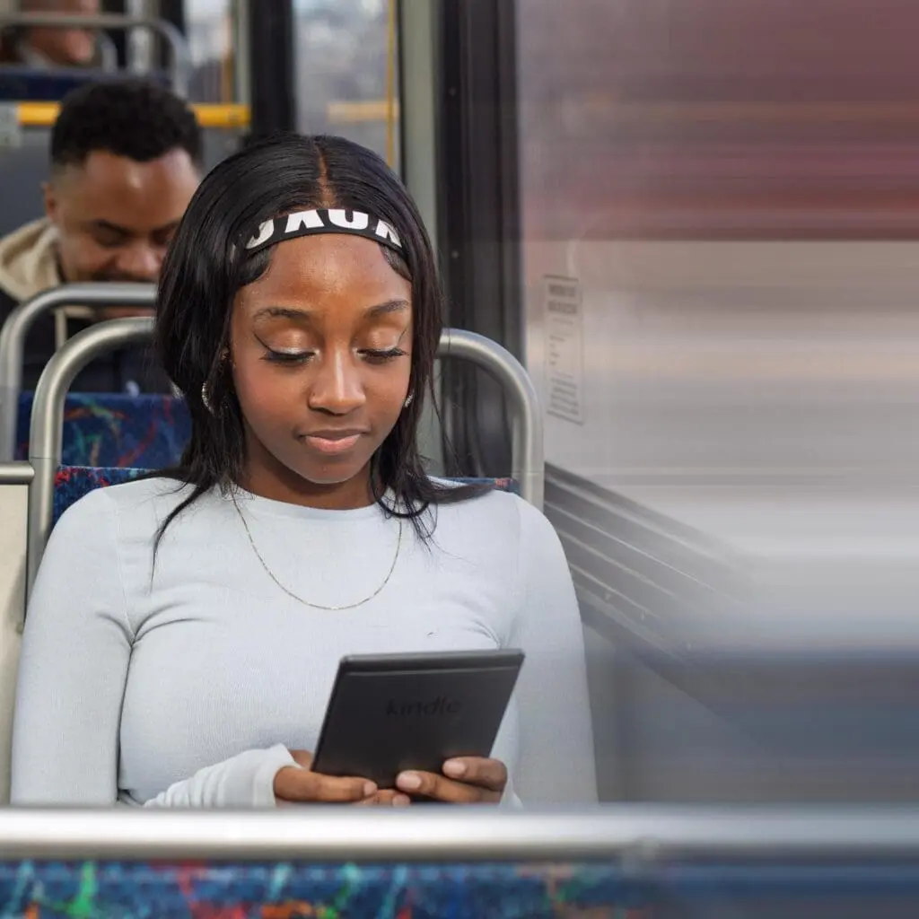 Young woman with a headband reading a Kindle e-reader while sitting on a bus.