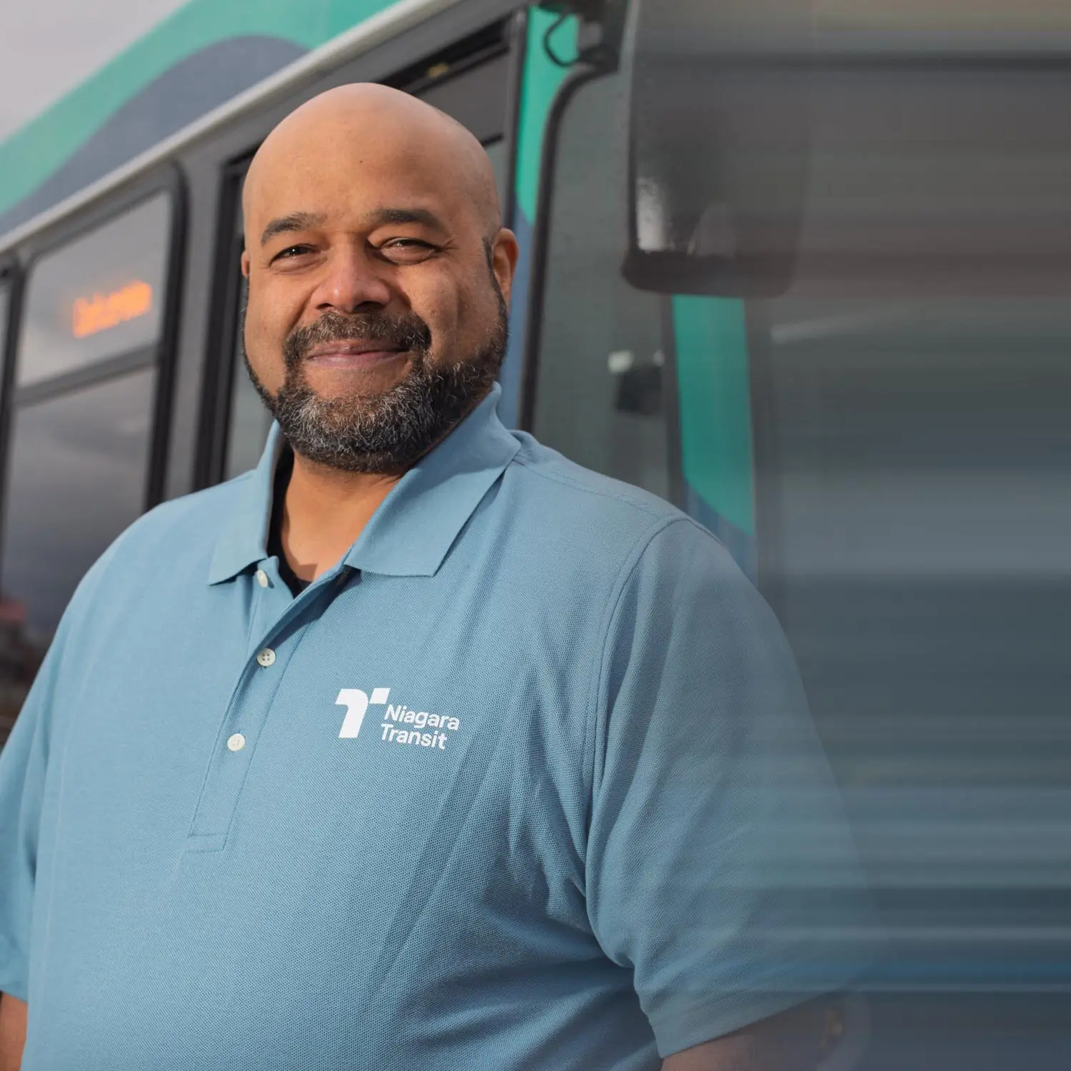 Smiling man wearing a light blue Niagara Transit polo shirt standing in front of a bus.