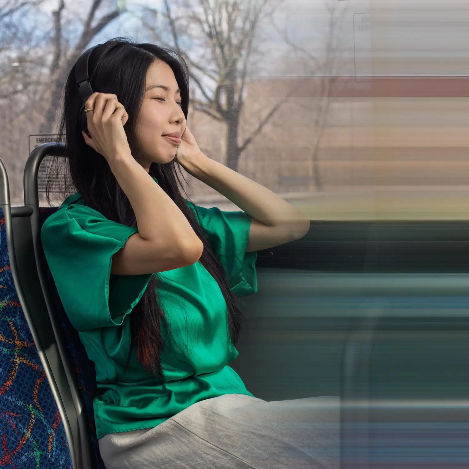 Woman in a green blouse wearing headphones and sitting on a bus with blurred motion outside the window.