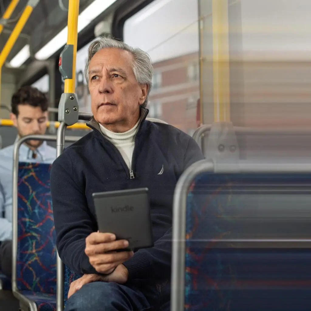 Older man with gray hair wearing a navy zip-up sweater and white turtleneck, sitting on a bus holding a Kindle e-reader, looking thoughtfully out the window.