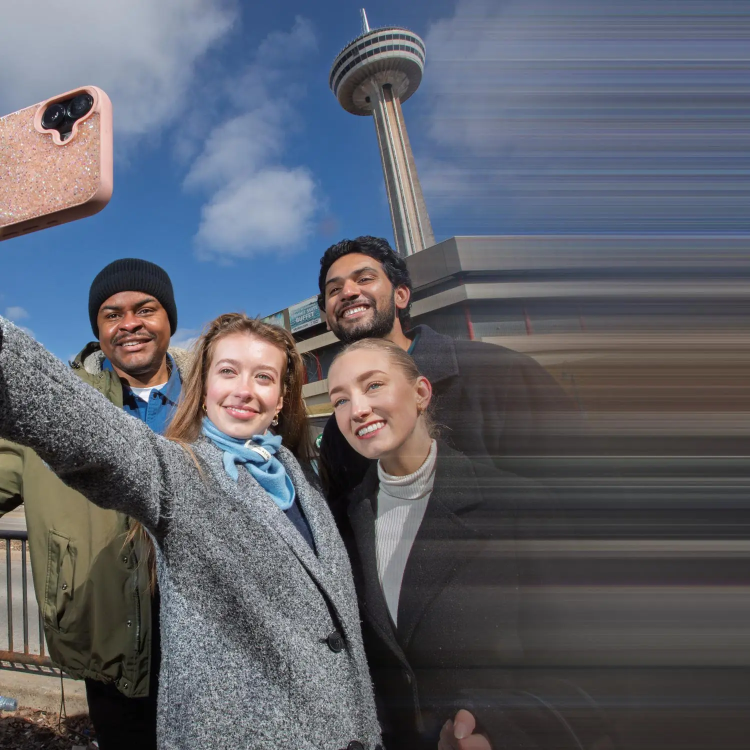 Four friends smiling and taking a selfie outdoors near a tall observation tower on a clear day.