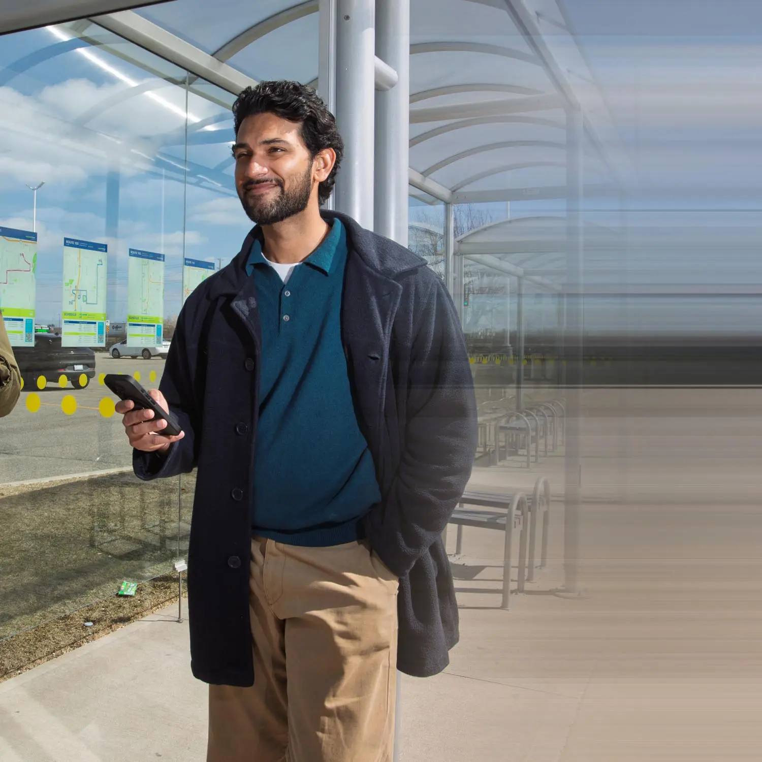 Man with dark hair and beard wearing a dark coat, teal shirt, and beige pants, holding a phone and standing at a bus stop with route maps on the glass behind him.