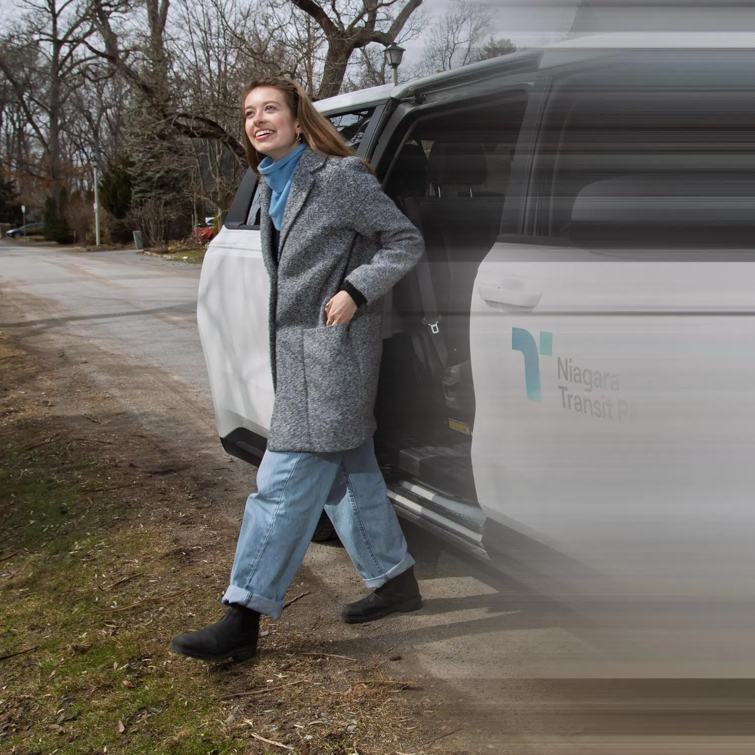 A smiling woman wearing a gray coat, blue scarf, light blue jeans, and black boots stepping out of a white Niagara Transit Plus vehicle on a roadside with bare trees in the background.