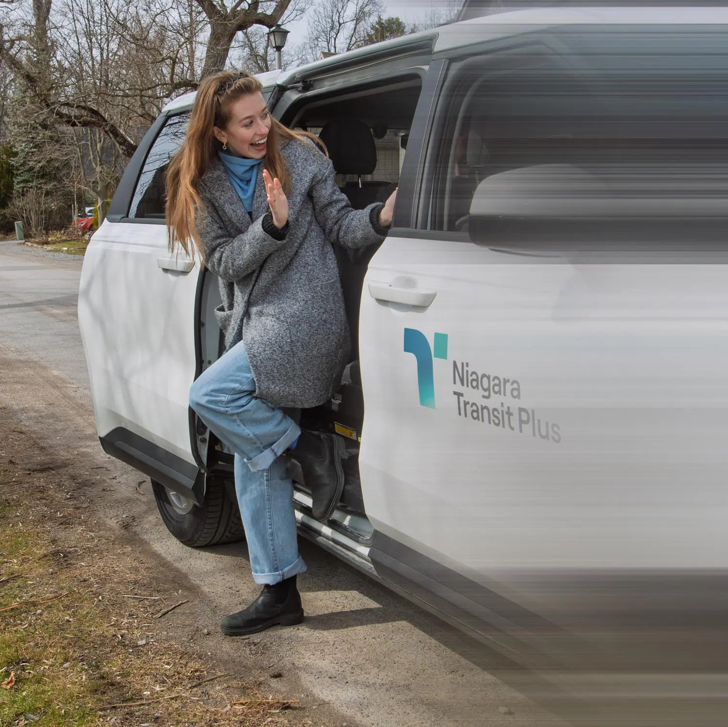 A woman wearing a gray coat, blue jeans, and black boots is stepping out of a white Niagara Transit Plus vehicle, smiling and waving.