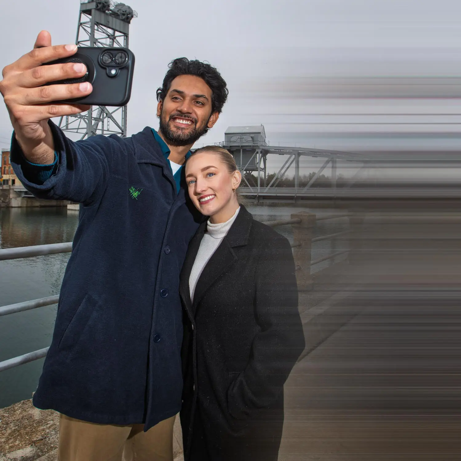 A smiling man and woman wearing coats taking a selfie near a waterfront with a metal bridge in the background.