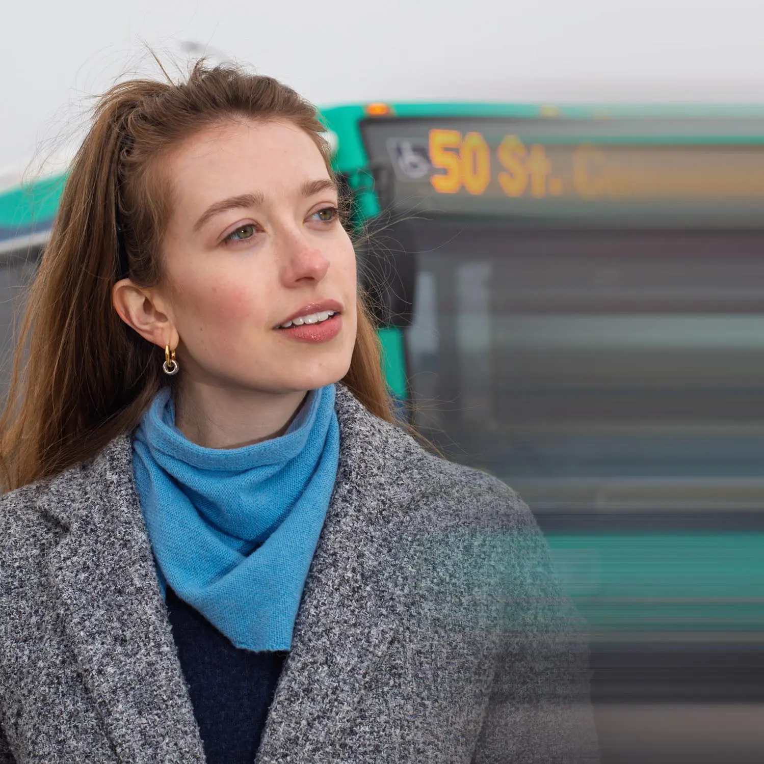 Young woman with long brown hair wearing a gray coat and blue scarf, standing in front of a green bus with a blurred destination sign.