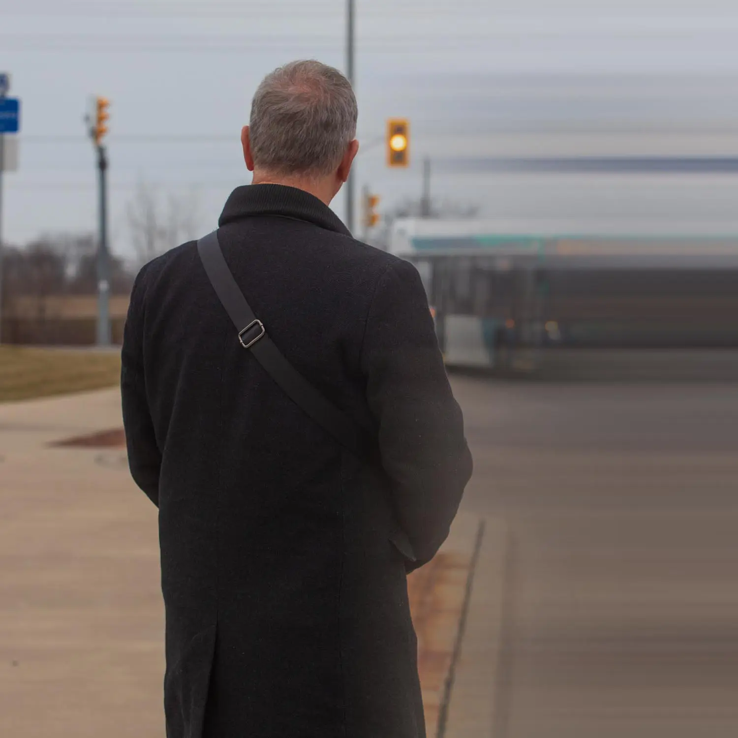 Man in a black coat with a shoulder strap standing on a sidewalk, facing away, with a blurred bus and traffic light in the background.