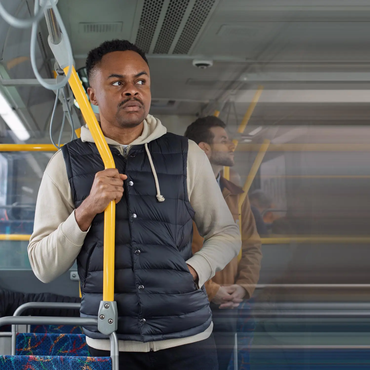 Man wearing a black puffer vest and beige hoodie standing and holding a yellow pole inside a bus.