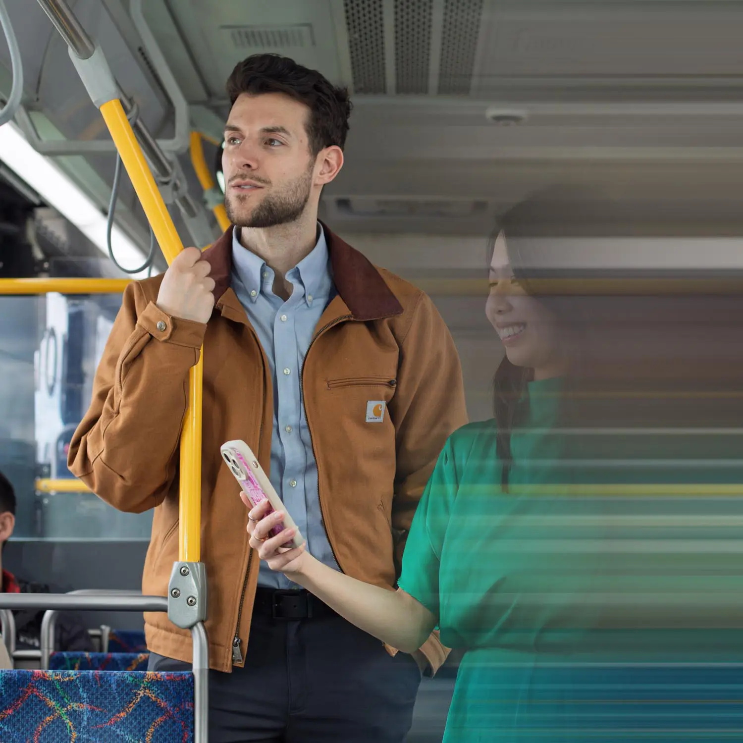 Man in a brown jacket holding a yellow pole on a bus, with a blurred woman in a green shirt holding a phone beside him.
