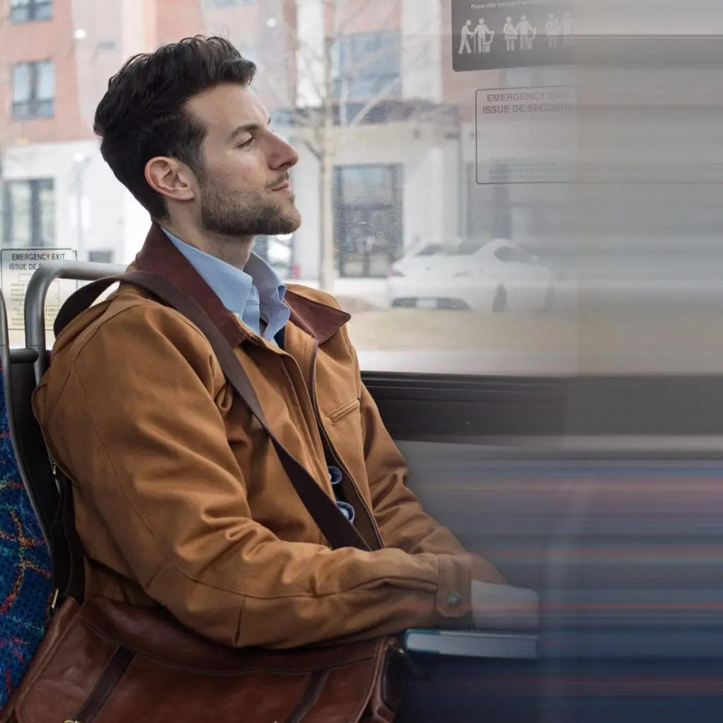 Man with a brown jacket and leather bag sitting on a bus, looking out the window.