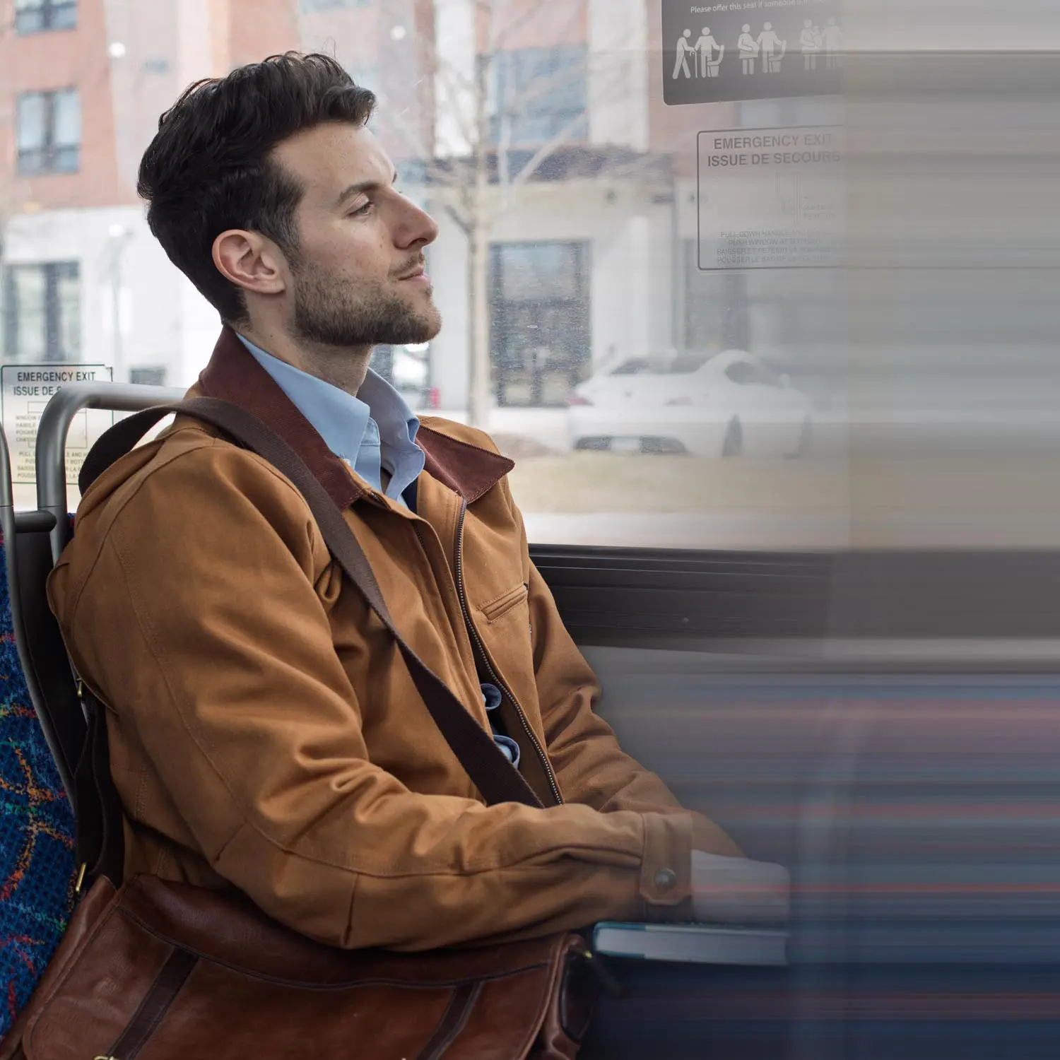 Man with a brown jacket and leather bag sitting on a bus, looking out the window.