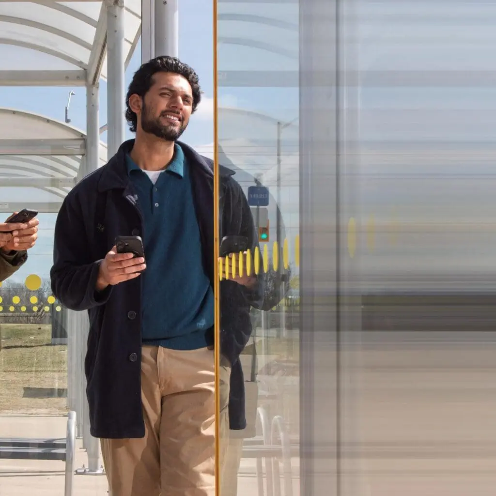 Man standing at a bus stop, holding a smartphone and smiling, with a glass panel partially obscuring his reflection.