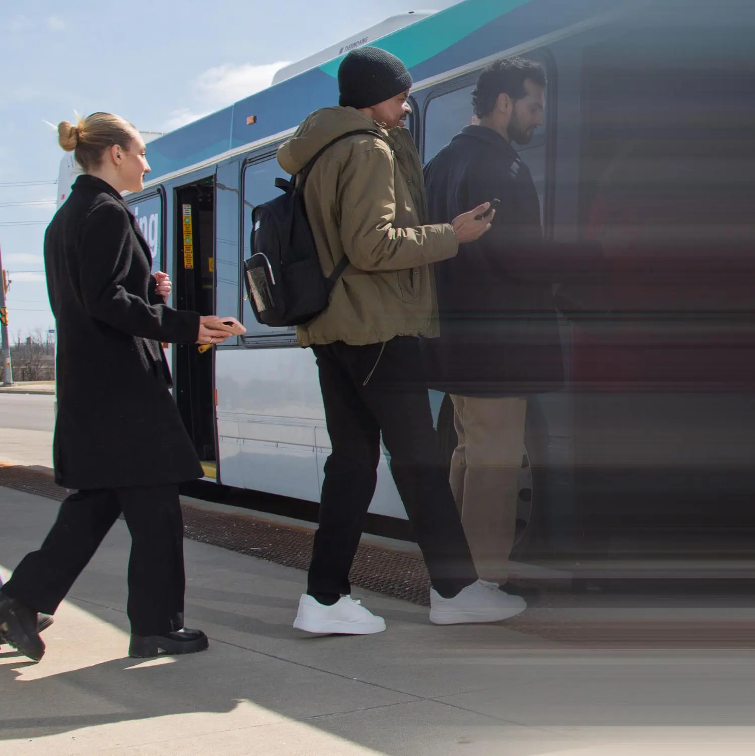 Three people boarding a city bus, with one person wearing a black coat, another in a green jacket and black beanie, and the third in a dark jacket and beige pants.