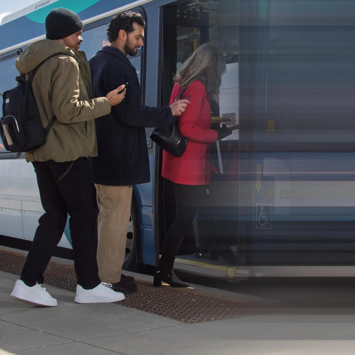Three people boarding a blue and white bus, with the person in front wearing a red coat and black boots.