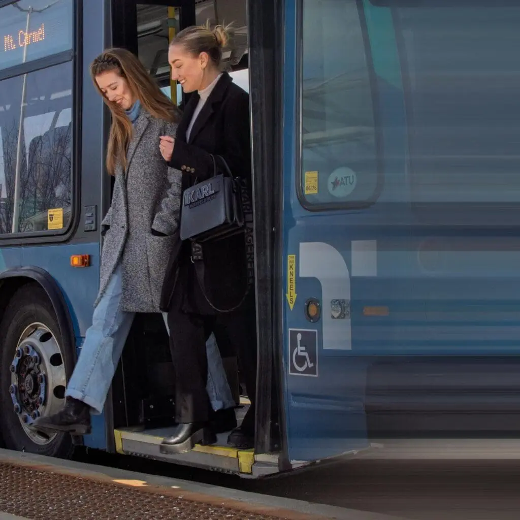 Two women stepping off a blue public bus, one wearing a gray coat and jeans, the other in a black coat holding a black handbag.
