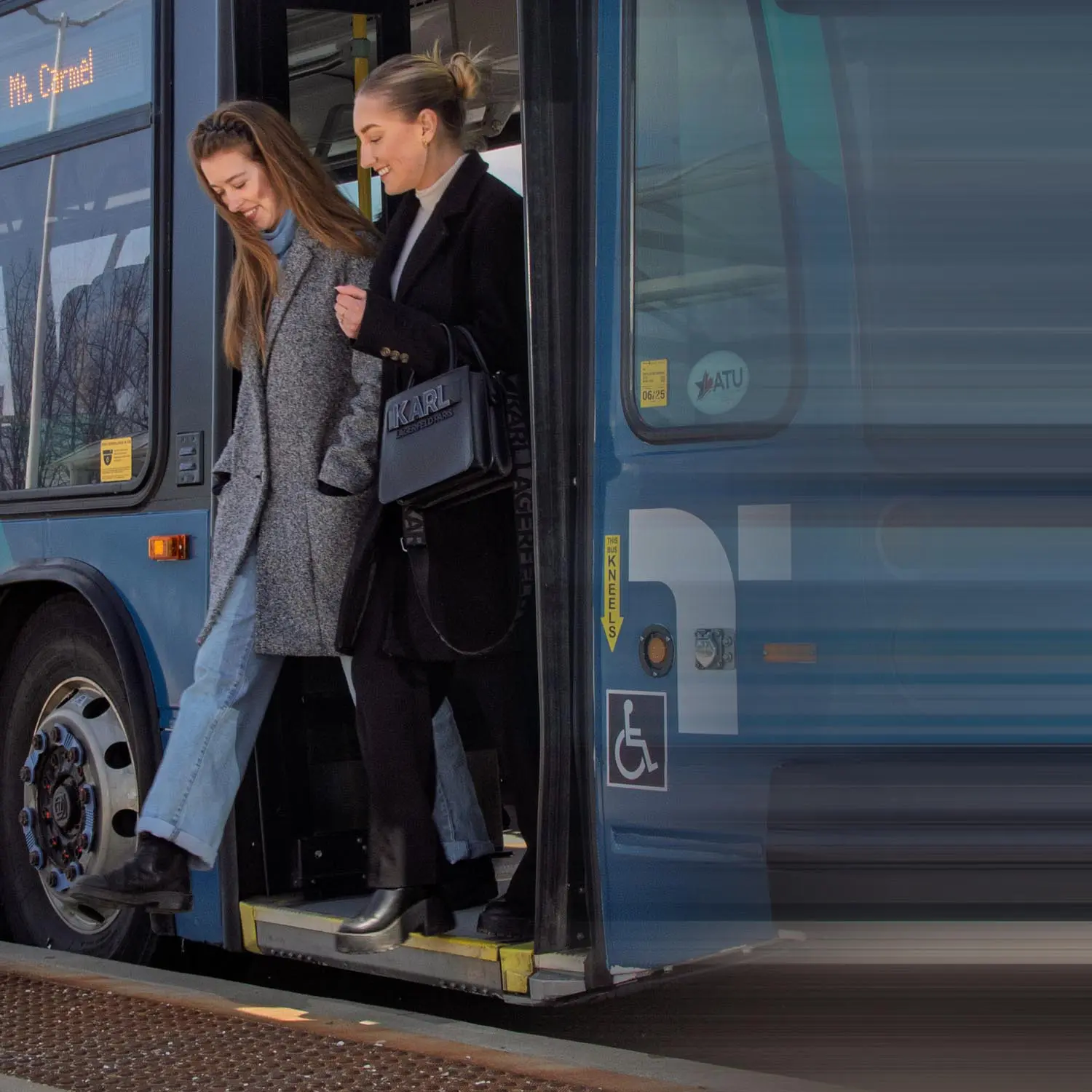 Two women stepping off a blue public bus, one wearing a gray coat and jeans, the other in a black coat holding a black handbag.
