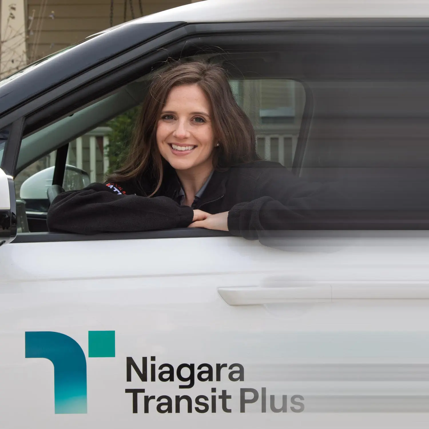 Smiling woman leaning out of the driver's side window of a white Niagara Transit Plus vehicle.