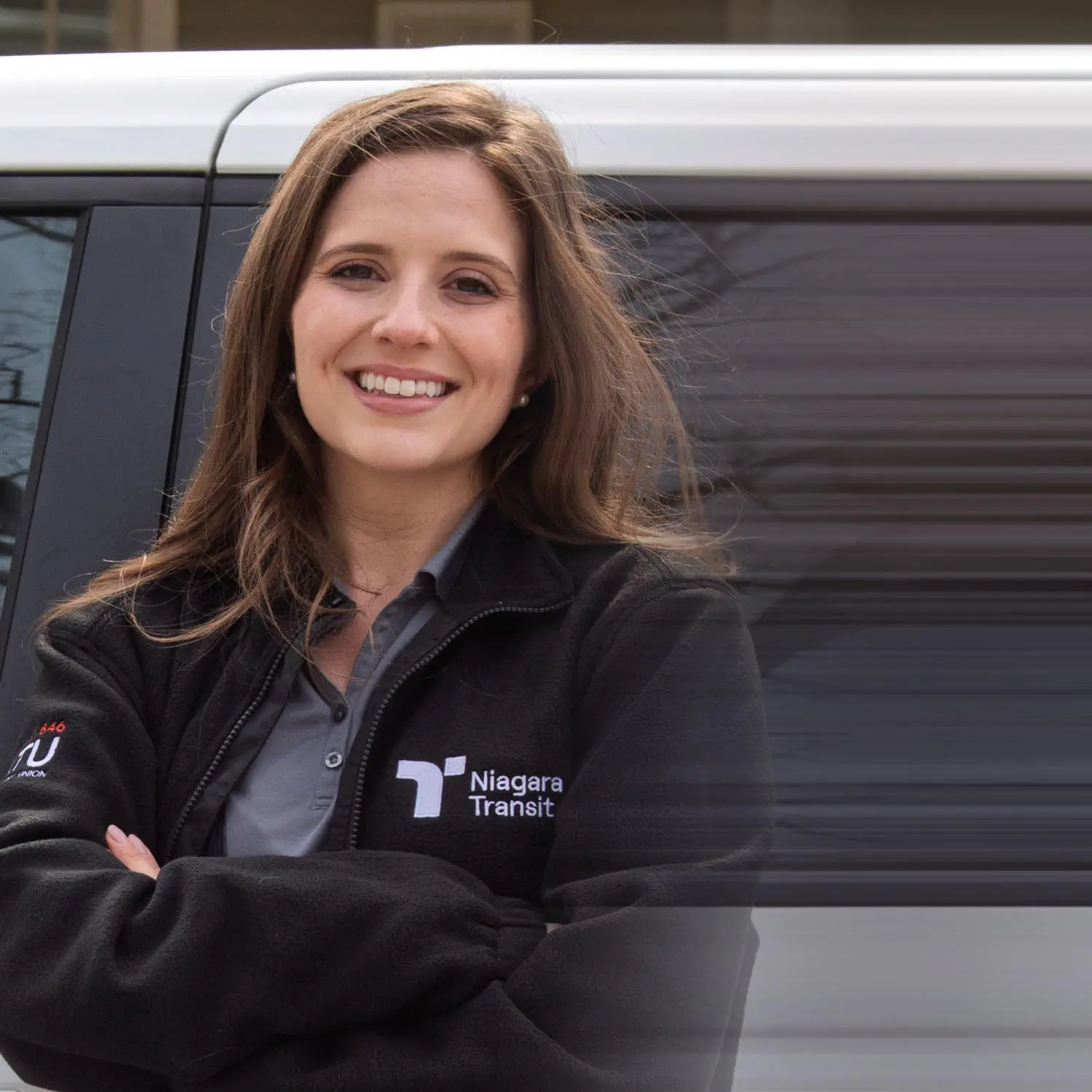 Smiling woman with long brown hair wearing a black Niagara Transit jacket, standing with arms crossed in front of a vehicle.
