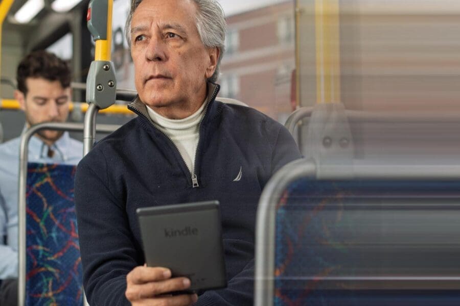 Older man with gray hair wearing a navy zip-up sweater and white turtleneck, sitting on a bus holding a Kindle e-reader, looking thoughtfully out the window.
