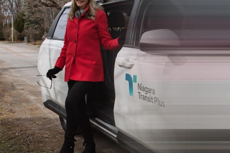 Woman in a red coat and black gloves standing next to an open door of a white Niagara Transit Plus vehicle.
