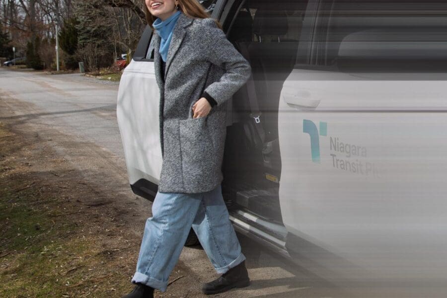 A smiling woman wearing a gray coat, blue scarf, light blue jeans, and black boots stepping out of a white Niagara Transit Plus vehicle on a roadside with bare trees in the background.