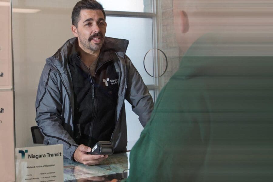 A man in a gray jacket and black Niagara Transit fleece is speaking to a customer across a glass service window, holding a handheld device.