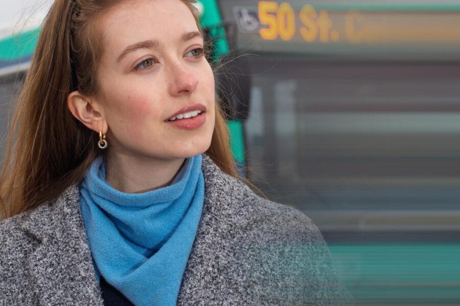 Young woman with long brown hair wearing a gray coat and blue scarf, standing in front of a green bus with a blurred destination sign.