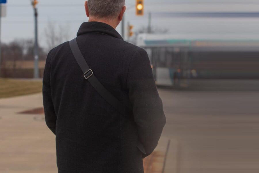 Man in a black coat with a shoulder strap standing on a sidewalk, facing away, with a blurred bus and traffic light in the background.