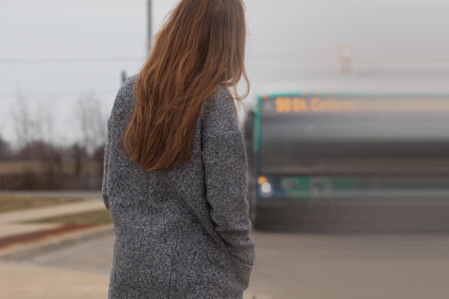 Woman with long brown hair wearing a gray coat standing on a sidewalk facing an approaching bus.