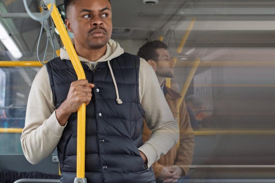 Man wearing a black puffer vest and beige hoodie standing and holding a yellow pole inside a bus.
