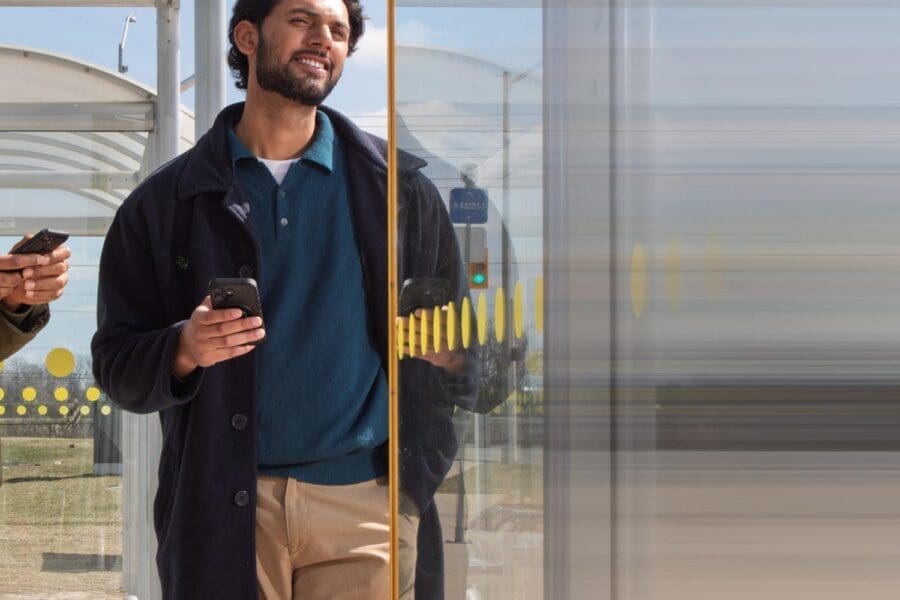 Man standing at a bus stop, holding a smartphone and smiling, with a glass panel partially obscuring his reflection.