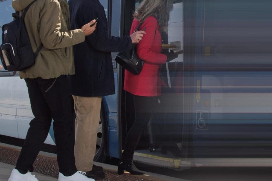 Three people boarding a blue and white bus, with the person in front wearing a red coat and black boots.