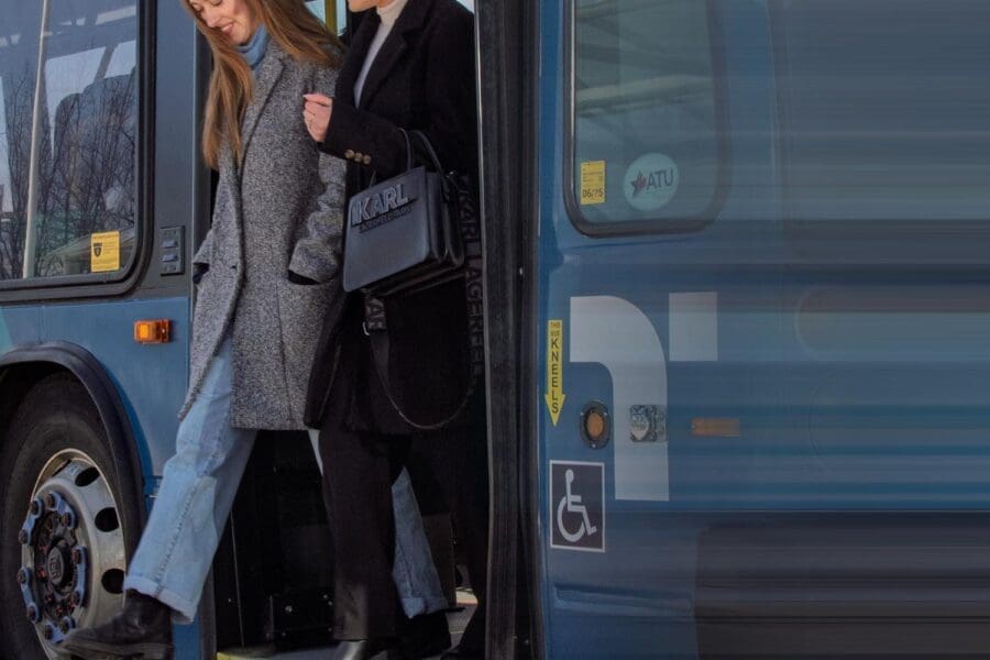 Two women stepping off a blue public bus, one wearing a gray coat and jeans, the other in a black coat holding a black handbag.