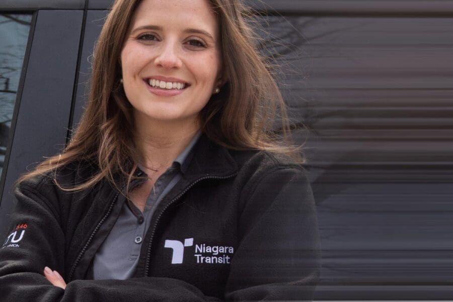 Smiling woman with long brown hair wearing a black Niagara Transit jacket, standing with arms crossed in front of a vehicle.
